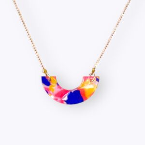 goudkleurige ketting met een hanger. De hanger is een halve cirkel met een vlekken patroon in de kleuren geel, fel roze, donker blauw en doorzichtig
